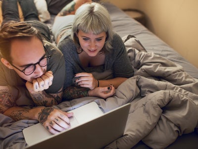 Ein Pärchen liegt auf dem Bett und schaut in einen Laptop.