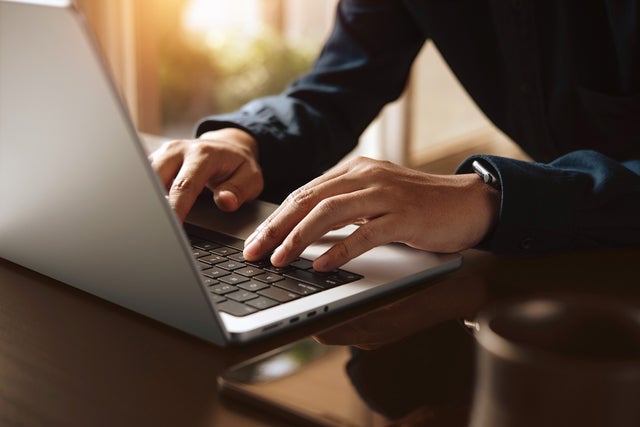 The picture shows a section of an open laptop from an angle so that only half of the keyboard is visible. A person in a blue shirt, of whom only the hands and arms can be seen, is in the right-hand half of the picture and is typing on the laptop keyboard. The hands and the laptop keyboard are in the centre of the picture and are in focus. To the right of the laptop, a smartphone and a black cup can be seen in a blur. In the background is a window that floods the room with light.