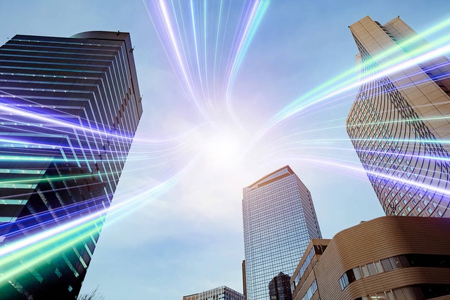 The image showcases a dynamic urban scene featuring several modern skyscrapers reaching towards the sky. The buildings are a mix of glass and steel structures, reflecting a professional and corporate environment. Superimposed over the scene are vibrant, multicolored light streaks that converge at a bright point in the sky, symbolizing connectivity and technology. The light streaks add a futuristic and energetic element to the image, suggesting advanced communication networks or data transfer within a bustling cityscape. The overall composition highlights the intersection of modern architecture and innovative technology.