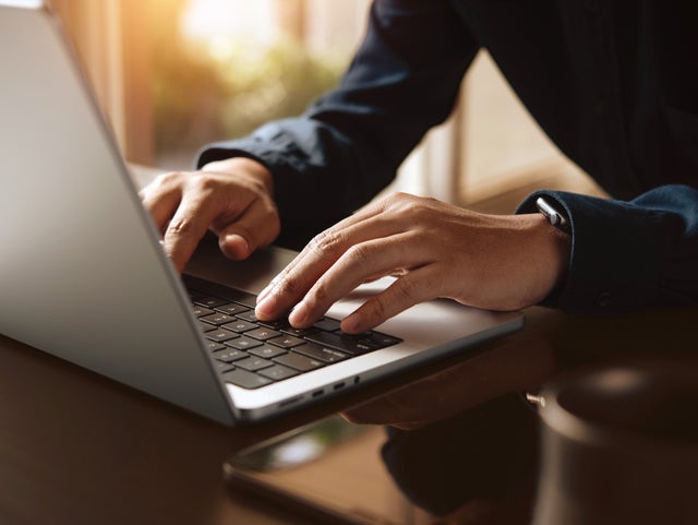 The picture shows a close-up of the keyboard of a laptop, which is illuminated by warm rays of sunlight from the background. The screen of the laptop is on the left edge of the picture and a woman in a dark blue shirt can be seen sitting in a blur on the right edge. Her hands are resting lightly on the keys of the laptop. The background of the picture is very blurred, but you can easily recognise the walls of a room and a window through which the sun's rays shine.