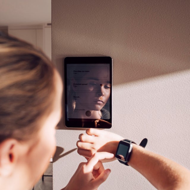 The image shows a person interacting with a tablet mounted on the wall. The person is seen from behind, with their reflection visible on the tablet screen. They are also wearing a smartwatch. The setting appears to be indoors, with natural light casting a shadow on the wall. The overall theme of the image emphasizes the integration of technology into everyday life, highlighting convenience and connectivity.