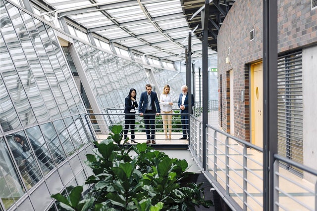 The picture shows four people in formal dress standing in a modern, light-flooded building. They are standing on a raised walkway with railings, surrounded by large glass windows. The windows extend along the entire length of the corridor and let in plenty of daylight.on the left of the picture is a woman with long, dark hair wearing a black trouser suit. She has her arms crossed in front of her chest and is looking down as if she is pensive. Next to her is a man in a dark blue suit, who is leaning on the railing with both hands and also looking down. Next to him is a woman with blonde hair in a white blazer and beige trousers. To her right is an older man with white hair and a beard, wearing a dark blue suit and interlocking fingers. The four people are discussing ista's building management system. Below the catwalk are large green plants that lend the room a natural element. On the right of the picture you can see a wall with dark red bricks and several doors. The whole space has a modern and open feel, with lots of natural light and a mix of industrial and natural elements.