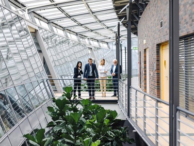 The picture shows four people in formal dress standing in a modern, light-flooded building. They are standing on a raised walkway with railings, surrounded by large glass windows. The windows extend along the entire length of the corridor and let in plenty of daylight.on the left of the picture is a woman with long, dark hair wearing a black trouser suit. She has her arms crossed in front of her chest and is looking down as if she is pensive. Next to her is a man in a dark blue suit, who is leaning on the railing with both hands and also looking down. Next to him is a woman with blonde hair in a white blazer and beige trousers. To her right is an older man with white hair and a beard, wearing a dark blue suit and interlocking fingers. The four people are discussing ista's building management system. Below the catwalk are large green plants that lend the room a natural element. On the right of the picture you can see a wall with dark red bricks and several doors. The whole space has a modern and open feel, with lots of natural light and a mix of industrial and natural elements.