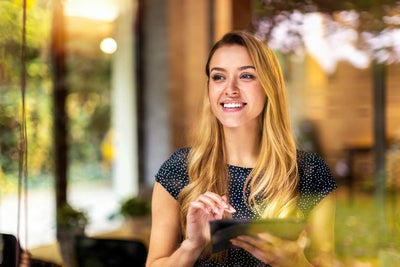 The picture shows a young woman looking through a window. She has long blonde hair and is wearing a dark blue, polka-dotted top. The woman is smiling and holding a tablet in both hands which is showing the Energy Audit Reporting from ista, her gaze directed slightly to the right. Green plants and a blurred light source can be seen in the background, giving the picture a warm and inviting atmosphere. The light source is located approximately in the centre of the background. The left background area shows a vertical black line, which is probably part of the window frame. The woman is standing relatively close to the window so that her reflection is partially visible in the glass. The foreground of the image is in focus while the background remains blurred, which draws the viewer's attention to the woman's smiling face and her tablet.