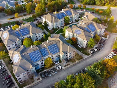 An aerial view of a residential complex showcasing several multi-story apartment buildings equipped with rooftop solar panels. The buildings are arranged in a circular layout, surrounded by well-maintained greenery, trees, and parking lots filled with vehicles. The roads curve around the complex, connecting the various structures. This image highlights the implementation of renewable energy through solar panels in a modern residential community, emphasizing sustainability and eco-friendly living practices. It illustrates the community's commitment to reducing its carbon footprint and embracing innovative energy solutions.