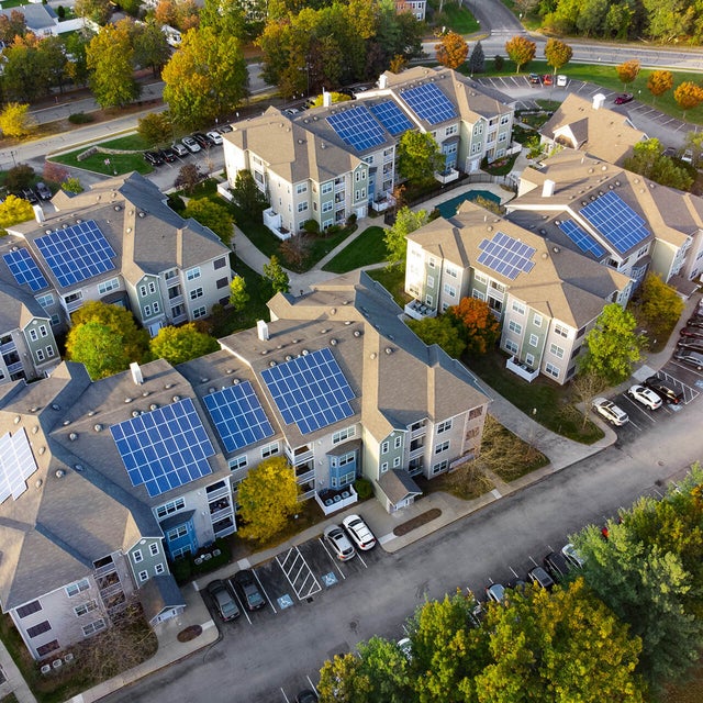 An aerial view of a residential complex showcasing several multi-story apartment buildings equipped with rooftop solar panels. The buildings are arranged in a circular layout, surrounded by well-maintained greenery, trees, and parking lots filled with vehicles. The roads curve around the complex, connecting the various structures. This image highlights the implementation of renewable energy through solar panels in a modern residential community, emphasizing sustainability and eco-friendly living practices. It illustrates the community's commitment to reducing its carbon footprint and embracing innovative energy solutions.