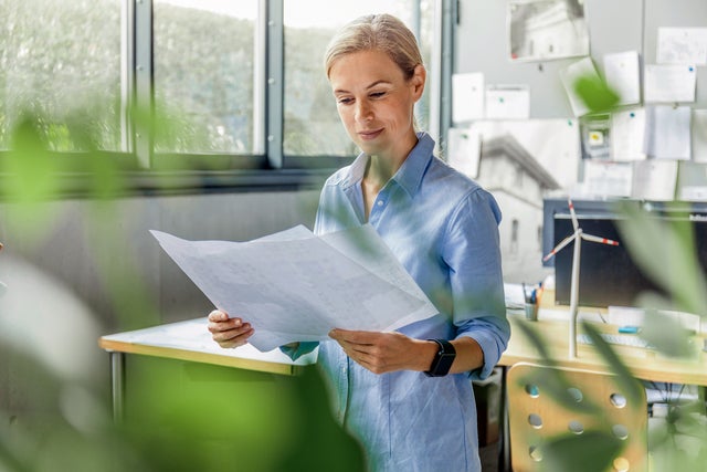 Durch die grünen Zweige einer Topfpflanze, sieht man eine Frau in einem Büro stehen. Sie steht mittig im Bild, trägt eine blaue Bluse und studiert Unterlagen, die Sie halbhoch in der Hand hält. Im Hintergrund ist ein Schreibtisch mit Bildschirm zu erkennen und eine Pinnwand.  