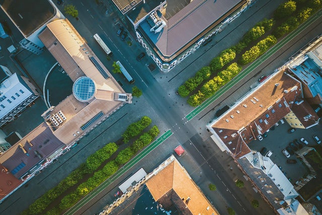 The picture shows a bird's eye view of the city. The picture was taken vertically downwards and shows an intersection from above, with two roads crossing in the centre of the picture. The road running from bottom left to top right is wider and appears to be a main road. It is lined with trees and a green strip on both sides that serves as a cycle path. The intersecting road, which runs from top left to bottom right, is slightly narrower and there are buildings with different roof shapes and colours at the four corners of the intersection. The building on the left has a striking, light brown roof with a round glass dome in the centre. Next to this building you can see a large bus parked on the street. The building at the top has a darker roof and is surrounded by several trees. The buildings on the right and below have red tiled roofs. The building on the right has a few cars in a car park next to it and the streets are quite clean with few vehicles or people to be seen. The green trees along the streets add a touch of nature to the urban environment. Overall, the picture appears calm and orderly, with clean lines and well-maintained urban structures.