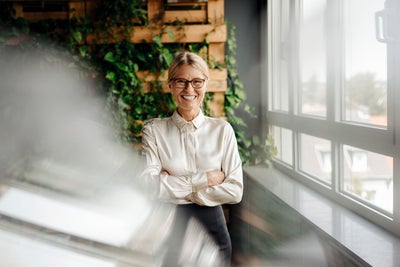 The picture shows a smiling woman with blonde hair and glasses standing in a modern office. She is wearing a cream-coloured blouse and has her arms crossed in front of her chest. The woman is in the centre of the picture. Behind her, a wall is covered with lush green foliage growing in a wooden frame. This green wall takes up the left part of the background and gives the room a natural and lively atmosphere. To the right of the woman, along the right side of the painting, is a window front that allows plenty of natural light into the room. The windows show a view of the roofs and the surroundings outside the building. A blur is visible in the foreground of the picture, possibly created by a glass surface or reflection. This blurring gives the image a certain depth and a modern photographic effect. The whole scene conveys an image of a modern, well-lit and green working environment that is both professional and inviting.