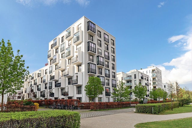 The picture shows a corner of a housing estate with white modern apartment buildings stretching from left to right across the picture. The buildings are characterised by an unusual architecture, as they alternately have protruding cubes with either large windows throughout the whole cube or set in balconies. The skyblue sky is filled with a few white clouds on the right side of the picture and in the foreground are red hedges and small trees that have been planted around the buildings, marking the property of the apartment buildings. There is a paved pedestrian path along the hedges and this is also bordered by another green hedge. The pedestrian path runs in a half circle in the bottom right of the picture, making a way through the grass that is planted there. 