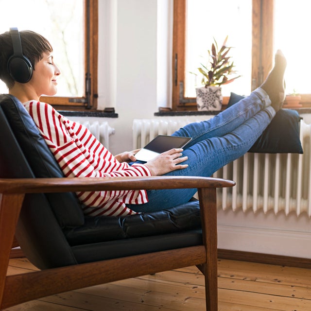 The picture shows a woman sitting comfortably on an armchair with wooden armrests and a black leather seat in front of a window. She has short brown hair and is wearing a white and red striped jumper and blue jeans. She is holding a tablet on her lap and listening to music through over-ear headphones. Her feet are propped up on a cushion resting on a radiator in front of the window. The floor of the room is made of light-coloured wood. On the windowsill, there is a flower pot with a green plant on the left and an empty white vase on the right.