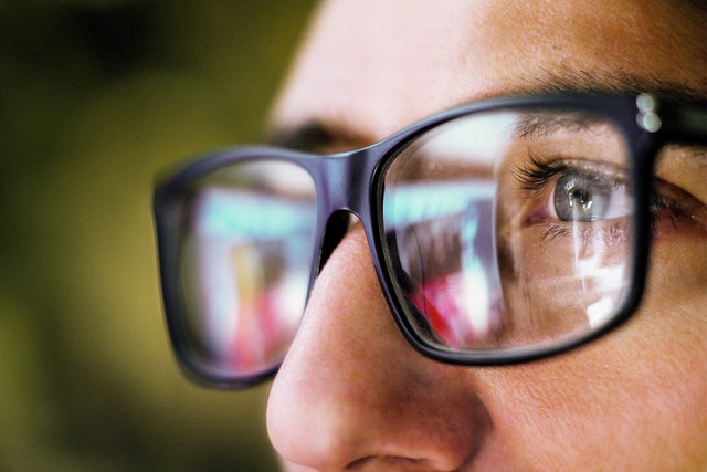 L'image montre un gros plan de la zone des yeux d'une personne portant des lunettes noires carrées. La personne regarde de la droite vers la gauche de l'image. On ne voit que l'œil gauche de la personne, qui est gris-bleu et entouré de cils foncés. L'accent est mis sur l'œil reconnaissable, le reste de l'image est flou.