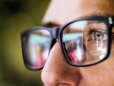 The picture shows a close-up of the eye area of a person wearing square, black glasses. The person is looking from the right side of the picture to the left. You can only see the person's left eye, which is grey-blue and surrounded by dark eyelashes. The focus of the picture is on the recognisable eye, the rest of the picture is blurry.