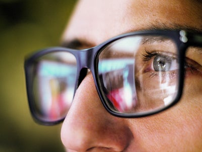 L'image montre un gros plan de la zone des yeux d'une personne portant des lunettes noires carrées. La personne regarde de la droite vers la gauche de l'image. On ne voit que l'œil gauche de la personne, qui est gris-bleu et entouré de cils foncés. L'accent est mis sur l'œil reconnaissable, le reste de l'image est flou.