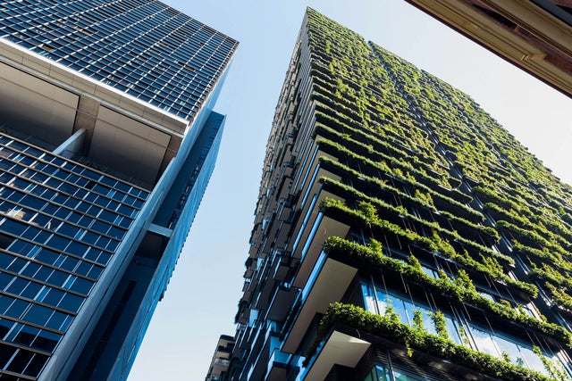 The picture shows a few skyscrapers that are seen from below. Two of them are clearly shown, the others are build behind them or can’t be seen from this point of view. On the skyscraper on the left side are just plain windows, but the skyscraper on the right side has greenery on every floor and is beeing illuminated by the sun.