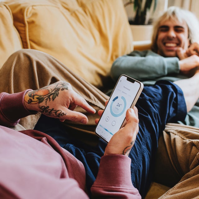 Man sieht zwei Männer auf der Couch. Der eine hält ein Smartphone in der Hand auf der eine App angezeigt wird.