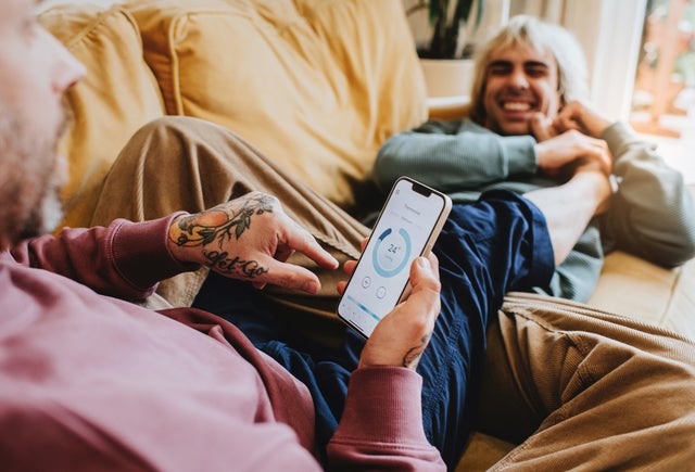 Man sieht zwei Männer auf der Couch. Der eine hält ein Smartphone in der Hand auf der eine App angezeigt wird.