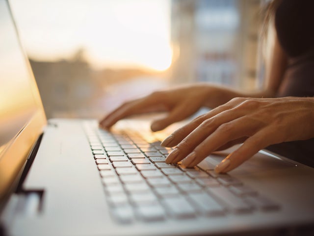 L'image montre un gros plan du clavier d'un ordinateur portable, qui est éclairé par des rayons de soleil chauds provenant de l'arrière-plan. L'écran de l'ordinateur portable se trouve sur le bord gauche de l'image et l'on peut voir une femme vêtue d'une chemise bleu foncé, assise dans un flou sur le bord droit. Ses mains reposent légèrement sur les touches de l'ordinateur portable. L'arrière-plan de l'image est très flou, mais on peut facilement reconnaître les murs d'une pièce et une fenêtre à travers laquelle passent les rayons du soleil.