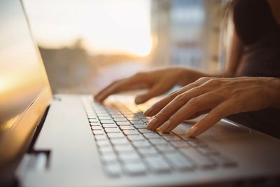 L'image montre un gros plan du clavier d'un ordinateur portable, qui est éclairé par des rayons de soleil chauds provenant de l'arrière-plan. L'écran de l'ordinateur portable se trouve sur le bord gauche de l'image et l'on peut voir une femme vêtue d'une chemise bleu foncé, assise dans un flou sur le bord droit. Ses mains reposent légèrement sur les touches de l'ordinateur portable. L'arrière-plan de l'image est très flou, mais on peut facilement reconnaître les murs d'une pièce et une fenêtre à travers laquelle passent les rayons du soleil.