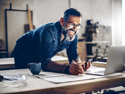 The picture shows a middle-aged man working in a workshop or studio. He has dark hair, wears glasses and his face is framed by a full, well-groomed grey beard. He wears a dark blue denim shirt and smiles broadly.The man is leaning over a large work table while holding a smartphone in his left hand looking at ista’s operating cost billing  and making notes on the paper with his right hand. There is an open laptop on the table in front of him. Next to the laptop is a blue coffee cup.The background of the workshop is blurred, but you can recognise shelves and tools that indicate a manual or creative activity. The light streaming in from the right through a window illuminates the scene and creates a warm, inviting atmosphere.The man appears focussed and content, which is conveyed by his smile and relaxed posture.