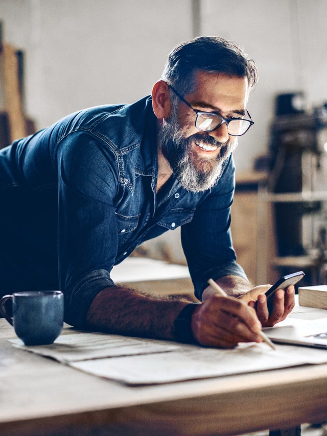 The picture shows a middle-aged man working in a workshop or studio. He has dark hair, wears glasses and his face is framed by a full, well-groomed grey beard. He wears a dark blue denim shirt and smiles broadly.The man is leaning over a large work table while holding a smartphone in his left hand looking at ista’s operating cost billing  and making notes on the paper with his right hand. There is an open laptop on the table in front of him. Next to the laptop is a blue coffee cup.The background of the workshop is blurred, but you can recognise shelves and tools that indicate a manual or creative activity. The light streaming in from the right through a window illuminates the scene and creates a warm, inviting atmosphere.The man appears focussed and content, which is conveyed by his smile and relaxed posture.