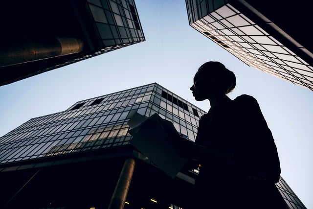 L'image montre la silhouette d'une femme tenant des documents sur fond de gratte-ciel modernes. La perspective de l'image est ascendante, ce qui donne aux bâtiments un aspect monumental et impressionnant. La femme se trouve au premier plan de l'image, légèrement tournée vers la gauche. Son profil est clairement visible et elle semble regarder attentivement ses documents. Elle porte une veste et a attaché ses cheveux en chignon. Plusieurs bâtiments en verre sont visibles à l'arrière-plan, occupant la majeure partie de la photo. Le bâtiment situé à gauche de l'image n'est que partiellement visible, avec un grand pilier rond s'élevant vers le haut. Le bâtiment central est le plus dominant et s'étend presque jusqu'en haut de l'image. Sa façade de verre et d'acier reflète le ciel. Un autre bâtiment est visible sur la droite de l'image, qui s'étend également vers le haut et forme un angle droit avec le bâtiment central. Les façades en verre des bâtiments donnent à l'image un aspect moderne et urbain. Le ciel est clair et légèrement bleuté, ce qui indique une journée ensoleillée ou au moins claire. 