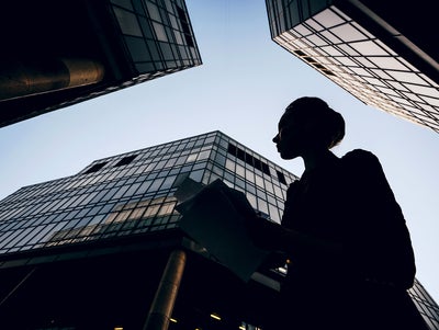 The picture shows a silhouette of a woman holding documents against the backdrop of modern skyscrapers. The perspective of the picture is from the bottom up, making the buildings look monumental and impressive. The woman is in the foreground of the picture, turned slightly to the left. Her profile is clearly visible and she appears to be looking intently at her documents. She is wearing a jacket and has tied her hair up in a bun. Several glass buildings can be seen in the background, taking up most of the picture. The building on the left of the picture is only partially visible, with a large round pillar extending upwards. The centre building is the most dominant and extends almost to the top of the picture. It has a glass and steel façade and reflects the sky. Another building can be seen on the right of the picture, which also extends upwards and forms a right angle to the centre building. The glass façades of the buildings give the picture a modern and urban feel. The sky is clear and slightly bluish, indicating a sunny or at least clear day. 