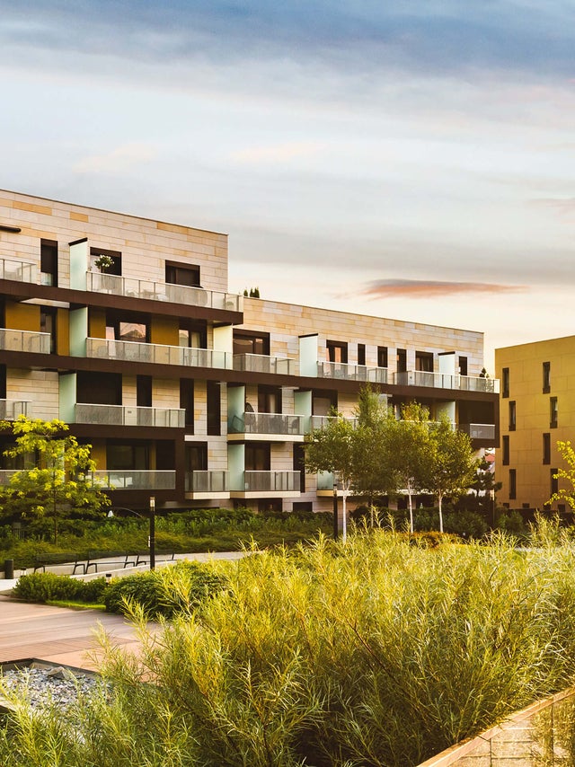 The picture shows a residential complex in the centre, stretching from the left to the right edge of the picture. The various buildings look very similar on the outside and have a white-orange brick façade. The individual flats have balconies with glass railings. The balconies are separated from each other by frosted glass walls and are furnished differently, giving each flat an individual touch. Most of the visible buildings have four floors, but vary in width so that different numbers of flats are visible on each floor. In the right quarter of the picture, in contrast to the others, another building rises up, which has the same style as the other buildings in the residential complex. A park can be seen in front of the residential complex in the lower half of the picture. Grasses and bushes surround the residential complex, accompanied by a pavement adorned with scattered trees. There are also benches and lanterns on the pavement. An artificial pond can be seen in the bottom left-hand corner, surrounded by a wooden walkway with glass railings. The sunset can be seen on the right-hand edge of the picture, giving the sky a slight colour gradient, from a reddish tone on the right to a bluish tone on the left.