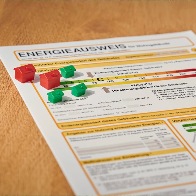 Ein Energieausweis liegt auf einer braunen Tischplatte. Auf dem Energieausweis stehen drei kleine, grüne und zwei etwas größere, rote Plastikhäuser.
