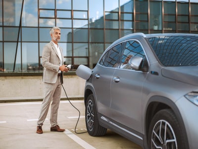 L'image montre une scène dans un parking moderne. Un homme âgé aux cheveux et à la barbe grisonnants, portant un costume gris clair et des chaussures marron, se tient à gauche de l'image. Il tient une prise de recharge dans sa main droite et regarde légèrement vers la droite en direction de la voiture électrique, qui se trouve à droite de l'image. La voiture électrique grise est représentée de côté, avec la trappe de chargement légèrement ouverte pour rendre le port de chargement accessible. L'homme semble être en train de recharger la voiture. Les grandes baies vitrées d'un bâtiment moderne sont visibles à l'arrière-plan, reflétant le paysage et donnant à l'image un aspect urbain et contemporain. Le sol du parking est plat et lumineux, et il n'y a pas d'autres véhicules ou personnes visibles, ce qui attire l'attention sur l'homme et la voiture.