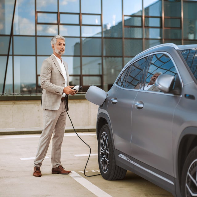 The picture shows an electric car that is being charged. In the foreground is the car's charging socket, into which a charging cable is plugged. The cable is connected to an ista EV charging station, which is outside the picture. In the background, slightly out of focus, is a man in a light-coloured suit talking on the phone. He has a relaxed posture and is leaning slightly against the car. The scene probably takes place in an urban environment or in a company car park, which is indicated by the modern buildings in the background. The image conveys an atmosphere of modern technology and environmentally friendly mobility.