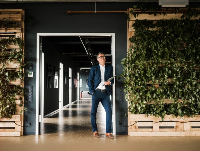 The picture shows a man with short grey hair and glasses in focus. He is wearing a blue suit with a white shirt and brown shoes. While his right hand is in his trouser pocket, he is holding a closed laptop in his other hand. He is leaning against a door frame that leads to a corridor in the background. Several doors can be seen on the left-hand side of the corridor, indicating various offices. To the right and left of the man on the walls are several vertically stacked pallets with green plants running down the pallets, giving the building a modern atmosphere.