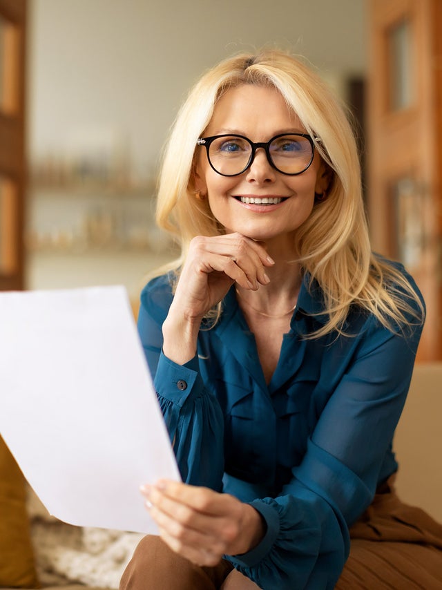 The picture shows a woman sitting on a couch and reading the ista heating bill. She has long blonde hair and is wearing black glasses. Her top is a blue shirt and she is wearing brown trousers. While she holds the heating bill in her left hand, her right hand rests on her chin. She looks directly into the camera. The beige couch on which she is sitting stretches across the entire lower third of the picture and has a white blanket draped over the back on the left with a yellow cushion leaning against it. In the background are various pieces of furniture that suggest a living room: A floor lamp and a shelf can be seen on the right, and in the centre an open two-part door leading into another room with bookshelves. The entire background is blurred.