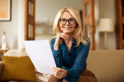 The picture shows a woman sitting on a couch and reading the ista heating bill. She has long blonde hair and is wearing black glasses. Her top is a blue shirt and she is wearing brown trousers. While she holds the heating bill in her left hand, her right hand rests on her chin. She looks directly into the camera. The beige couch on which she is sitting stretches across the entire lower third of the picture and has a white blanket draped over the back on the left with a yellow cushion leaning against it. In the background are various pieces of furniture that suggest a living room: A floor lamp and a shelf can be seen on the right, and in the centre an open two-part door leading into another room with bookshelves. The entire background is blurred.