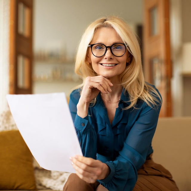 The picture shows a woman sitting on a couch and reading the ista heating bill. She has long blonde hair and is wearing black glasses. Her top is a blue shirt and she is wearing brown trousers. While she holds the heating bill in her left hand, her right hand rests on her chin. She looks directly into the camera. The beige couch on which she is sitting stretches across the entire lower third of the picture and has a white blanket draped over the back on the left with a yellow cushion leaning against it. In the background are various pieces of furniture that suggest a living room: A floor lamp and a shelf can be seen on the right, and in the centre an open two-part door leading into another room with bookshelves. The entire background is blurred.