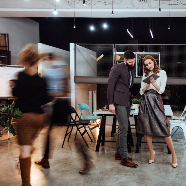 The picture shows a modern, open-plan office space where two people are standing in the center of the room. In the background on the left are cupboards containing files and a presentation board. To the right, you can see two white walls raised close together that do not reach the ceiling of the room. A railing is attached to the right-hand wall, which extends to the right-hand side of the picture and to the wall there. The wall of the railing itself is made of a grey grid and the floor is attached to a steel beam that can be seen from the side. The lattice provides a clear view of the black wall behind it. Two large desks can be seen in the center of this elevation, with two chairs on either side. On the right-hand side, against the brick wall, there is another desk with a chair and a passageway in the wall. In the foreground you can see two people walking towards the cupboards on the left. These two people are only vaguely recognizable as they are in motion and therefore blurry. In front of the railing in the center of the picture are two large tables that have been placed opposite each other and form a large group of tables. There are two chairs to the left and right of the group of tables. Three plants can be seen on the right-hand edge of the picture, their branches protruding into the picture. In the center of the picture, a younger man is standing next to a younger woman, right before the group of tables. The man has short black hair and a black beard and is wearing a black jacket with grey trousers and brown shoes. The woman has open red wavy hair and is wearing a white blouse, a grey metallic skirt and white pumps. They are both smiling while the woman has a tablet in her hand and is presenting the smart metering options from ista to the man.