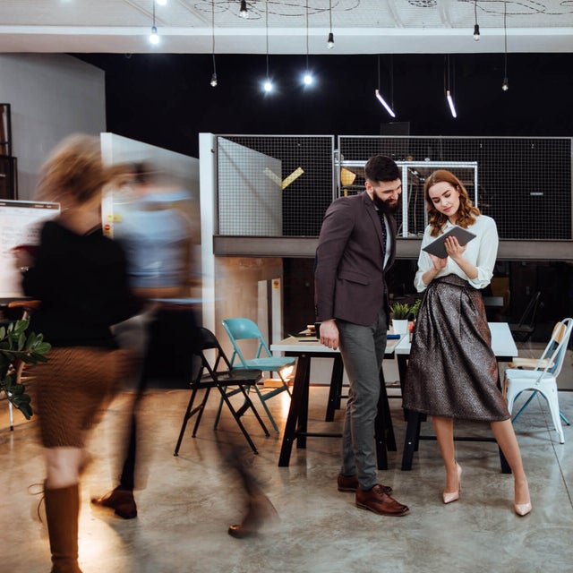 The picture shows a modern, open-plan office space where two people are standing in the center of the room. In the background on the left are cupboards containing files and a presentation board. To the right, you can see two white walls raised close together that do not reach the ceiling of the room. A railing is attached to the right-hand wall, which extends to the right-hand side of the picture and to the wall there. The wall of the railing itself is made of a grey grid and the floor is attached to a steel beam that can be seen from the side. The lattice provides a clear view of the black wall behind it. Two large desks can be seen in the center of this elevation, with two chairs on either side. On the right-hand side, against the brick wall, there is another desk with a chair and a passageway in the wall. In the foreground you can see two people walking towards the cupboards on the left. These two people are only vaguely recognizable as they are in motion and therefore blurry. In front of the railing in the center of the picture are two large tables that have been placed opposite each other and form a large group of tables. There are two chairs to the left and right of the group of tables. Three plants can be seen on the right-hand edge of the picture, their branches protruding into the picture. In the center of the picture, a younger man is standing next to a younger woman, right before the group of tables. The man has short black hair and a black beard and is wearing a black jacket with grey trousers and brown shoes. The woman has open red wavy hair and is wearing a white blouse, a grey metallic skirt and white pumps. They are both smiling while the woman has a tablet in her hand and is presenting the smart metering options from ista to the man.