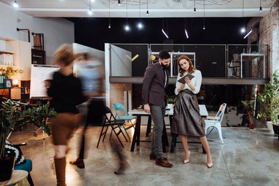 The picture shows a modern, open-plan office space where two people are standing in the center of the room. In the background on the left are cupboards containing files and a presentation board. To the right, you can see two white walls raised close together that do not reach the ceiling of the room. A railing is attached to the right-hand wall, which extends to the right-hand side of the picture and to the wall there. The wall of the railing itself is made of a grey grid and the floor is attached to a steel beam that can be seen from the side. The lattice provides a clear view of the black wall behind it. Two large desks can be seen in the center of this elevation, with two chairs on either side. On the right-hand side, against the brick wall, there is another desk with a chair and a passageway in the wall. In the foreground you can see two people walking towards the cupboards on the left. These two people are only vaguely recognizable as they are in motion and therefore blurry. In front of the railing in the center of the picture are two large tables that have been placed opposite each other and form a large group of tables. There are two chairs to the left and right of the group of tables. Three plants can be seen on the right-hand edge of the picture, their branches protruding into the picture. In the center of the picture, a younger man is standing next to a younger woman, right before the group of tables. The man has short black hair and a black beard and is wearing a black jacket with grey trousers and brown shoes. The woman has open red wavy hair and is wearing a white blouse, a grey metallic skirt and white pumps. They are both smiling while the woman has a tablet in her hand and is presenting the smart metering options from ista to the man.