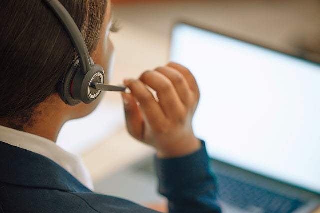 The picture shows a woman from the support center with a gray headset with a microphone sitting before an open laptop that is shining in a bright white. The picture was taken over the shoulder of the woman. The woman has her hair back and is wearing a white shirt and a dark blue jarkett and with er right hand she is adjusting the microphone of her headset.