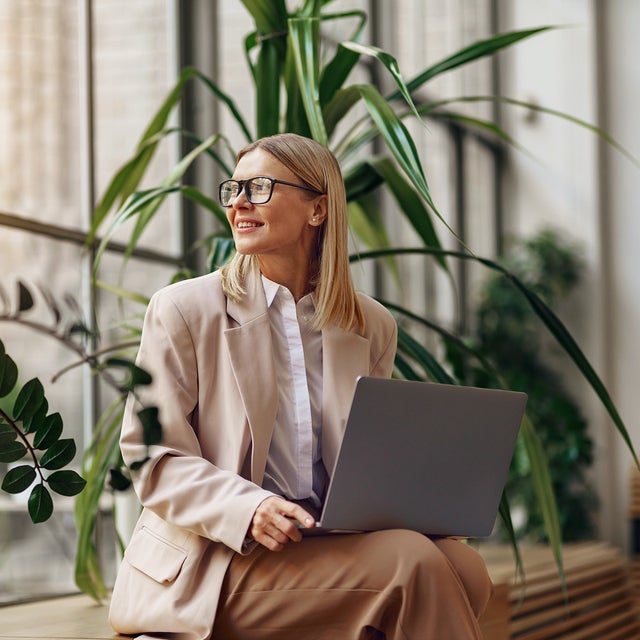 The image features a professional woman seated in a modern office setting, illuminated by natural light from large windows. She is wearing a beige blazer over a white blouse and matching beige pants. The woman has shoulder-length blonde hair and is wearing glasses. She is holding a laptop on her lap and looking outside with a pleasant and focused expression, because the energy accounting from ista is making everything easier. The background includes several lush green plants, adding a touch of nature to the workspace.