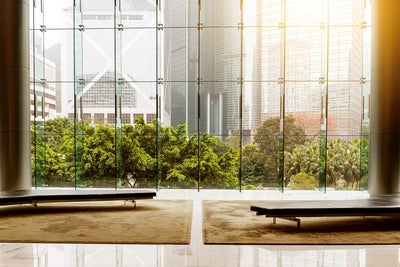 L'image montre un hall d'entrée ou une salle d'attente moderne avec une impressionnante façade vitrée qui offre une vue sur un paysage urbain. Au premier plan de l'image se trouvent deux bancs noirs simples posés sur une grande moquette beige. Les bancs sont disposés symétriquement l'un par rapport à l'autre, l'un à gauche et l'autre à droite, laissant un espace vide entre eux. Le sol est constitué de carreaux blancs polis qui reflètent l'environnement et la lumière entrante, créant ainsi une impression de clarté et de convivialité. Le mur de verre à l'arrière-plan se compose de plusieurs panneaux hauts et étroits qui s'élèvent jusqu'au plafond et sont reliés par des entretoises métalliques. Une végétation verte et luxuriante est visible à travers le mur de verre, contrastant avec les grands immeubles de bureaux modernes à l'arrière-plan. Les bâtiments ont des styles architecturaux différents et reflètent la lumière de différentes manières, ce qui donne de la profondeur et du dynamisme à la scène. Le côté gauche montre un bâtiment remarquable avec une structure diagonale caractéristique, tandis que le côté droit montre plusieurs immeubles de grande hauteur avec des façades en verre. Le soleil pénètre dans l'image en haut à droite, baignant la scène d'une lumière chaude et dorée.