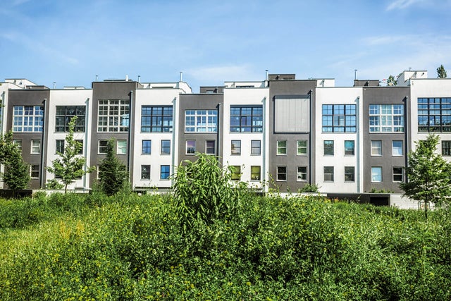 La photo montre une rangée de maisons mitoyennes étroites mais hautes. Les façades des maisons mitoyennes sont alternativement grises et blanches. A gauche, vous pouvez voir une maison avec une façade blanche et à l'extrême droite, une maison avec une façade grise. Elles sont également construites à des hauteurs différentes, de sorte qu'il n'y a jamais deux maisons de même hauteur l'une à côté de l'autre. Toutes les maisons ont une grande fenêtre carrée dans la moitié supérieure, composée de 30 petites fenêtres carrées. La façade de la quatrième maison en partant de la droite est la seule à être couverte par un store. Toutes les maisons ont quatre fenêtres individuelles sous les façades, deux à gauche et deux à droite l'une en dessous de l'autre. Au-dessus des maisons, on peut voir un ciel bleu presque entièrement dégagé. In the foreground in front of the houses you can see a lawn with many bushes and a total of four trees, three of which can be seen on the left at the edge of the picture and one tree on the right.