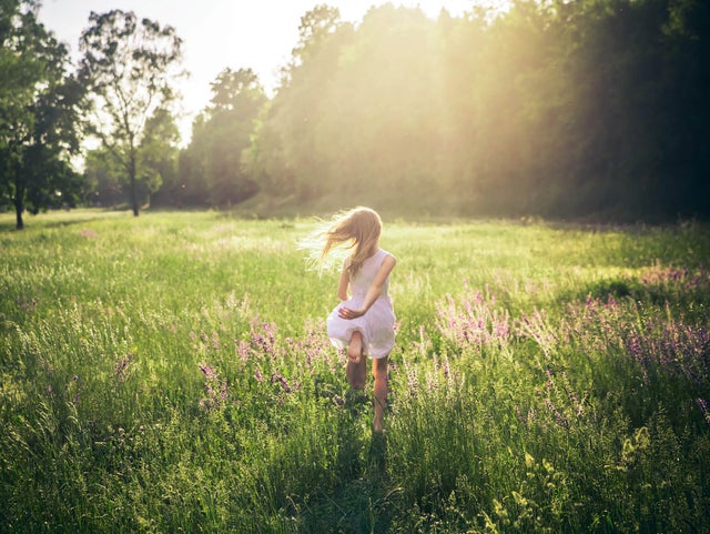 The picture shows a green, wide and tall meadow with large green trees on the edge of the horizon in the upper third of the picture and a girl running through the meadow. From the upper edge in the centre, bright yellow rays of sunlight stretch across the middle of the picture and bathe it in warm light. In the centre of the picture, a little girl in a knee-length, sleeveless white dress is walking across the meadow with her back to the viewer. Her long blonde hair blows away from her body to the left as she moves. In addition, as she runs, her left arm is loosely behind her back and her left foot is bent upwards so that the entire underside of her foot can be seen at the end of her dress.