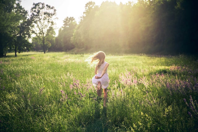 The picture shows a green, wide and tall meadow with large green trees on the edge of the horizon in the upper third of the picture and a girl running through the meadow. From the upper edge in the centre, bright yellow rays of sunlight stretch across the middle of the picture and bathe it in warm light. In the centre of the picture, a little girl in a knee-length, sleeveless white dress is walking across the meadow with her back to the viewer. Her long blonde hair blows away from her body to the left as she moves. In addition, as she runs, her left arm is loosely behind her back and her left foot is bent upwards so that the entire underside of her foot can be seen at the end of her dress.