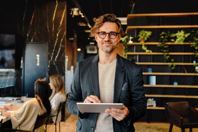 The picture shows a smiling man holding a tablet on which ista's Energy Management Minute View is displayed. The man has brown hair and is wearing black glasses. He is wearing a beige and grey striped shirt and a blue blazer over it. He is standing in the centre of the picture and looking directly into the camera. A modern office can be seen in the background. On the left-hand side of the picture is a wooden shelf in front of a black wall, decorated with long hanging plants and various objects that give the impression of awards. On the right of the picture, two women are sitting next to each other at a white work table covered with various work items. The woman in front, whose long black hair and beige loose top are visible, rests her arms on the table and has turned her head towards her colleague, so that only the back of her head is visible. The woman next to her has long blonde hair and is wearing a light blue shirt. Her side profile is recognisable. The entire background is blurred so that the focus is on the smiling man with the tablet.