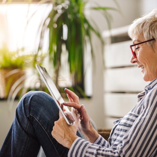 The picture shows a relaxed scene in which an elderly woman is sitting comfortably on the floor. She is turned sideways to the camera and leaning slightly backwards against a white cupboard. Her right arm is bent while she holds the tablet in both hands and touches the screen with the fingers of her right hand. The woman is wearing glasses with red frames and a blue and white striped shirt. Her hair is short and blonde and she smiles as she looks at the tablet. There are several green plants in pots in the background. The largest plant is at the top left of the picture, right next to a window. This window allows warm, natural light to stream into the room, creating a cosy and bright atmosphere. Further left in the picture, closer to the foreground, a smaller plant can be seen, which is slightly out of focus and contributes to the warm atmosphere. To the right of the woman, in the background, other blurred plants can be seen, giving the room a fresh and lively look. These plants are at different heights and distances from the camera, adding depth to the image. The woman is sitting in a slightly bent position, with her legs drawn up and crossed. The overall effect of the room is inviting and cosy, which is enhanced by the warm light and the green plants.