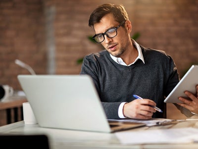 In the picture, a man wearing glasses and a gray sweater sits at a desk. On the desk, located in the foreground, are a laptop on the left and a tablet in his right hand. He is looking at the tablet while holding a pen in his left hand. In the background, there is a brick wall and blurred office items, including a mug and other objects on another desk to the left.