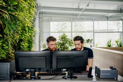 The picture shows a light-flooded part of an office space. Two men can be seen sitting at a desk directly in the foreground, stretching from left to right in the lower third of the picture. The man sitting on the left has short, dark brown and curly hair, a brown-red beard, wears round glasses with black frames and a black shirt. The man to his right has almost black short hair, a brownish beard and is wearing a black T-shirt. Both men are looking at the two monitors on the desk in front of them. Both men are looking at the carbon audit provided by ista on these monitors. To the left of the monitors on the desk is a black printer. To the right of the monitors is a telephone and next to it, on the right-hand edge, two letter trays are stacked. In the background you can see a wall consisting of large windows that stretches from the first left third of the picture to the right edge. In addition, the ceiling, which can be seen in the upper part of the picture, also consists of the same large windows. On the right-hand side of the picture, a dark brown, approximately waist-high room divider can be seen, which bends to the right once. Six plants can be seen on the room divider and behind it. To the left of the picture, directly next to the man on the left, is a wall consisting entirely of green plants.