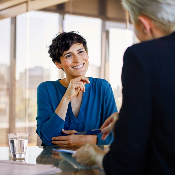 The picture shows two people sitting opposite each other at a table in a room filled with warm sunlight and with glass walls. On the left side of the table is a full glass of water and asmall pile of documents. The picture was taken from the perspective of one person, so that you only see the back of them and the other person, who is a woman, is in focus. The woman is in the center of the picture and the other person is on the right side of the picture, beeing a bit cut off. The woman is wearing a bright blue shirt, has dark wavy short hair and is smiling at the other person while holding her right hand to her chin and the other hand lying on the table touching her ellbow. The person that can only be seen from the back has a black jacket on and bright blond hair/ grey hair and a short sleek back haircut. They are explaining something to the woman while they are holding a document with their left hand and pointing with a ballpoint pen with their right hand onto something on the document.