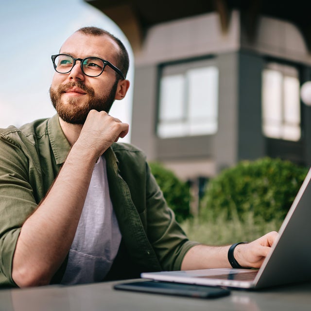 A young professional man, wearing glasses and a casual green jacket, is seated at an outdoor café with a laptop open in front of him. He appears thoughtful and focused, with his hand resting on his chin. On the table beside the laptop are a smartphone and a notepad. The background features modern office buildings and greenery. This image conveys a sense of contemplation and productivity, emphasizing the concept of consumption data monitoring in a business environment.